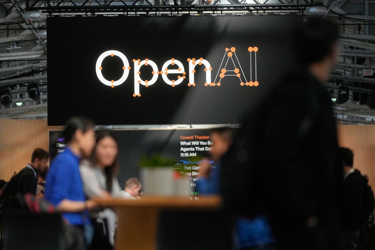 A large sign in a conference center reads "OpenAI" as people sit at a table or mill about in the foreground out of focus.