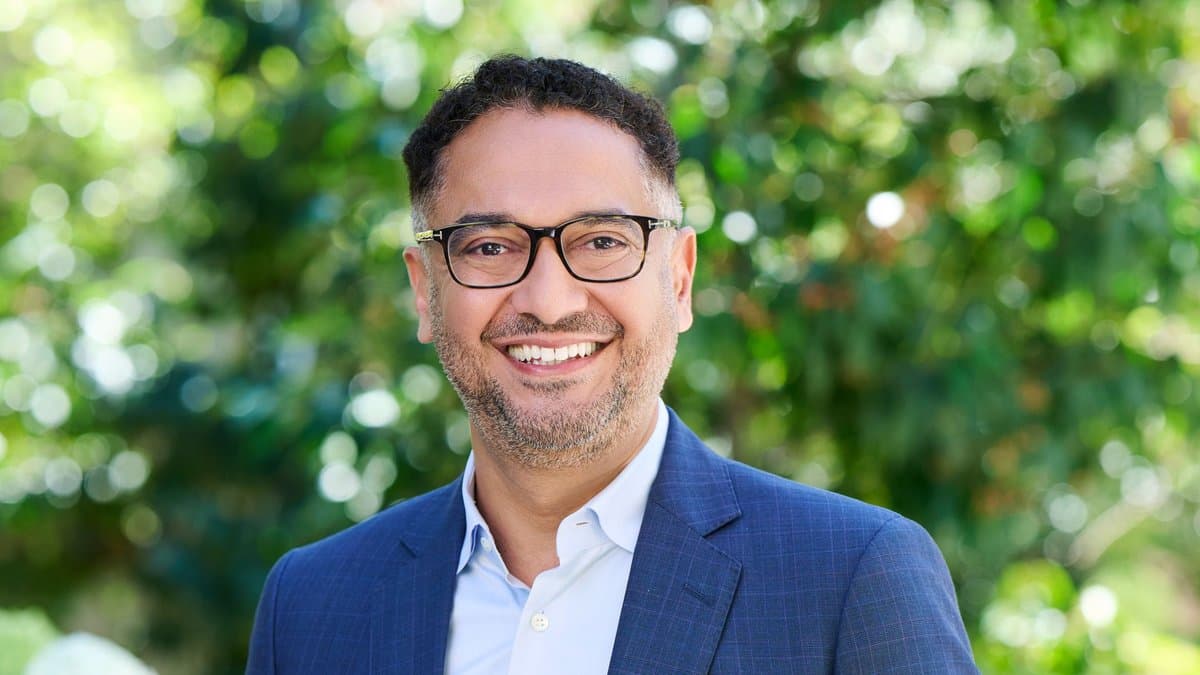 A man in a navy blue suit and light blue shirt smiles at the camera, wearing glasses, standing outdoors in a sunny garden with lush green foliage in the background.